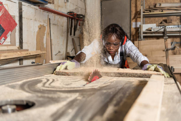 Photo of one African-American female carpenter using circular Female worker wearing ear and eye protectors while working. Carpenter is cutting wood with circular saw on workbench. She is making furniture in workshop. carpenter stock pictures, royalty-free photos & images