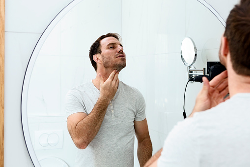 Reflection of man with beard looking at mirror and touching face in bathroom