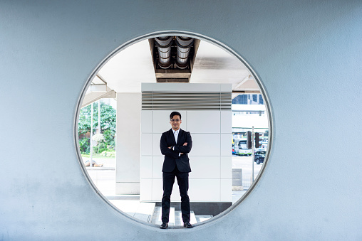 Full length front view of confident Chinese businessman wearing suit and standing in curve of oculus architectural feature with arms crossed smiling at camera.