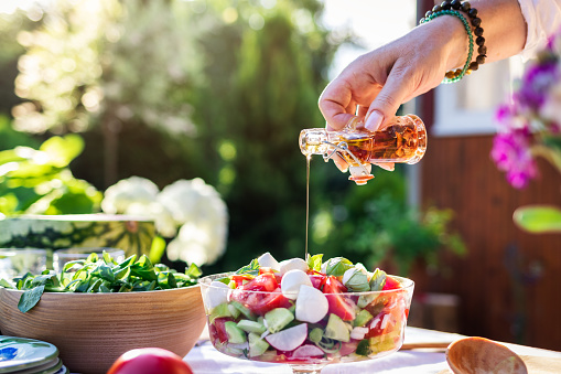 Woman hand pouring flavored olive oil to fresh vegetable salad. Preparation healthy vegetarian food for garden party outdoors