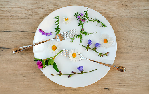 Grass and flowers with fork and spoon in a white plate on a wooden table, vegetarian food or diet concept.