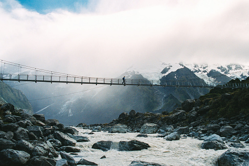 Female Hiker crossing a suspension bridge over a glacier river with snowcapped mountains and a glacier behind