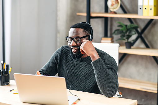 Clever young African-American guy wearing stylish eyeglasses with headset sits on his workplace in the modern office, he looks at laptop screen, smiles and talking online. Remote work, work on the distance