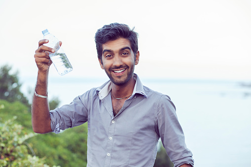 Hydrate yourself, you should drink more concept. Man smiling in profile about to drink from a bottle of water isolated nature trees and sea background