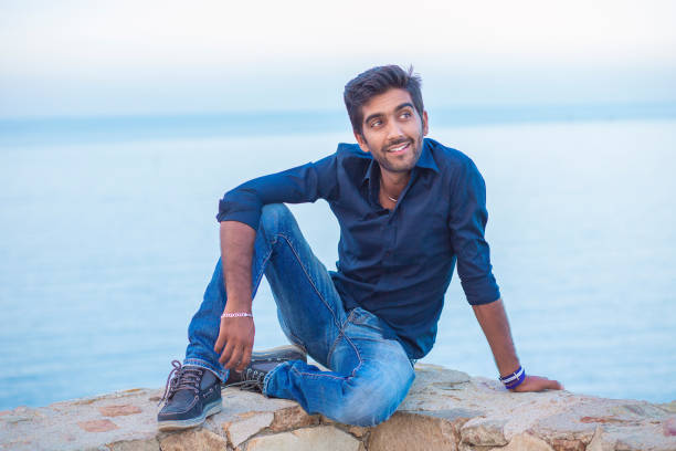 istock Man smiling looking at camera happy celebrating freedom while sitting on a concrete wall bridge above the sea. 1254584261