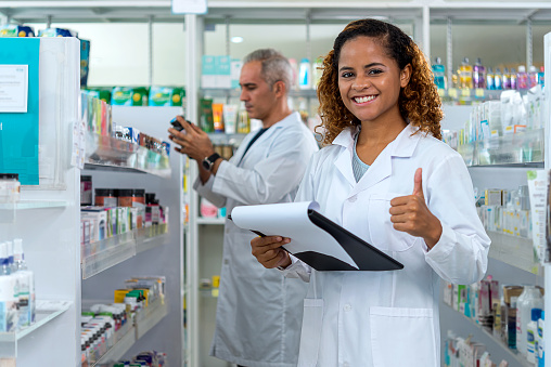 Portrait of a female pharmacist standing and holding clipboard with her  colleague in background.