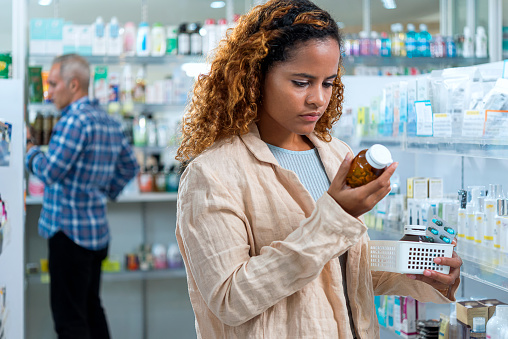 Young female customer holding pill container and read to medical instruction before decision to purchase. Background with another customer buying medicine.