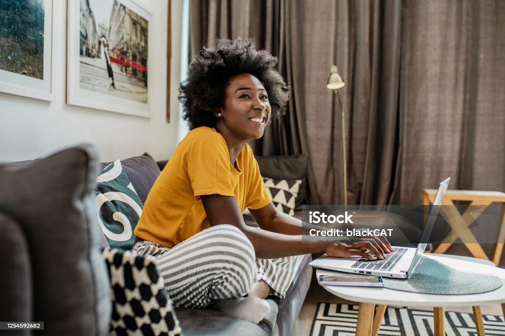 A young African-American woman uses a laptop in her modern apartment A young African woman is using a laptop and working in the living room while smiling and enjoying in a cozy environment Apartment Stock Photo