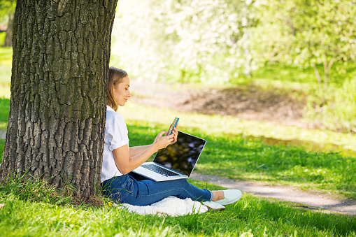 Young blond woman on grass close to tree in summer park using laptop,