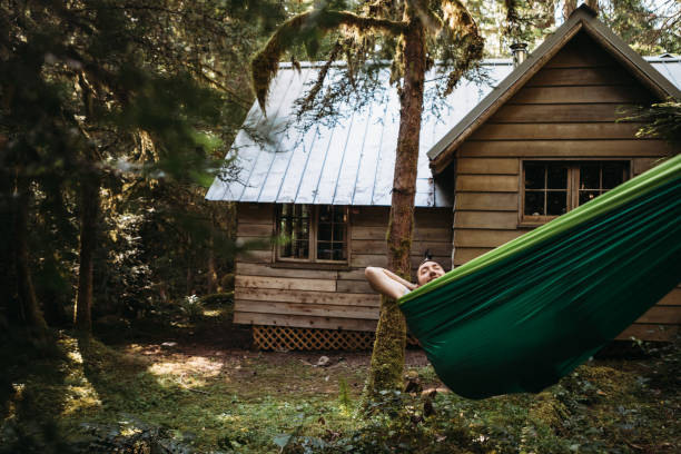 An adult Caucasian man relaxes in a camping hammock strung between two trees outside a beautiful isolated log cabin in the woods in the Pacific Northwest.  Shot in Washington state, USA.