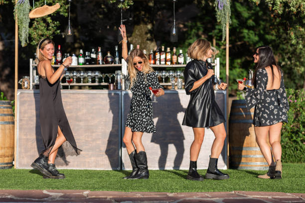 黒いパーティードレスを着た4人の白人の女の子が、夏の低い太陽で日中に屋外パブバーの前で踊ります - friendship relaxation cocktail nightclub ストックフォトと画像