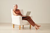 Middle-aged freelance woman in stylish outfit sitting into comfortable armchair and using laptop while working on online project