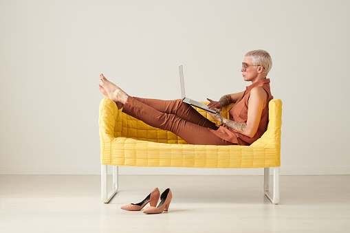 Serious relaxed business lady with short blond hair sitting on yellow sofa and surfing websites on laptop