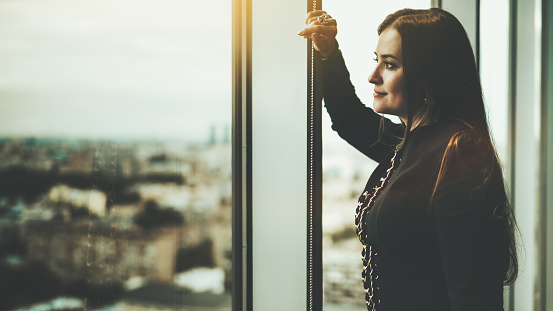 Side view of a charming caucasian woman leaning against the window frame and wistfully observing evening cityscape from a top floor of her apartment; a copy space place on the left for an ad message
