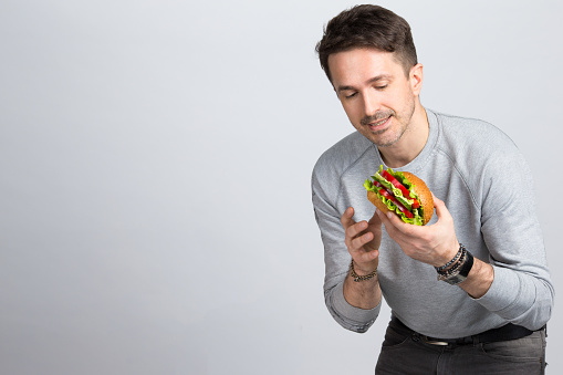 Handsome young modern man holding and looking at a big vegan burger a sandwich in your hands on gray background.
