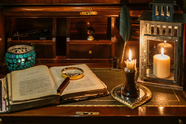 open antique book on which a magnifying glass rests. are found on a wooden desk on leather. two lighted lanterns are incorporated to illuminate with a candle and two other writing pens - cargill, incorporated imagens e fotografias de stock