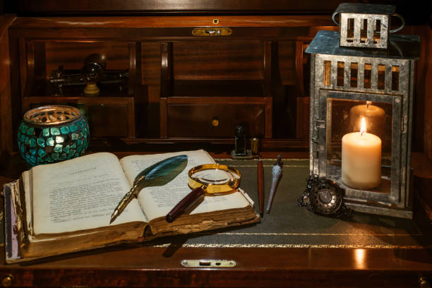 un libro antiguo abierto en el que descansan una lupa y un bolígrafo de escritura. se encuentran en un escritorio de madera sobre cuero. dos linternas encendidas se incorporan para iluminar con una vela - bible study university old fashioned fotografías e imágenes de stock