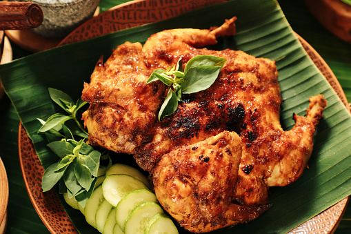 Ayam Bekakak, the traditional Sundanese grilled chicken dish. Spices-marinated whole chicken is grilled on charcoal fire. It is plated on a ceramic plate lined with fresh cucumber slices and lemon basil leaves.