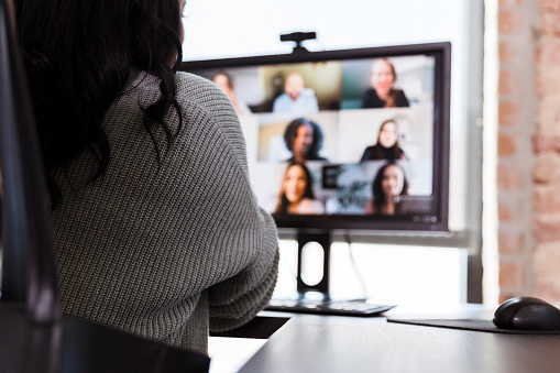 Rear view of a businesswoman on a video conference with a group of colleagues. Focus is on the woman, the computer monitor is blurred.