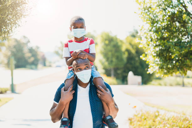 Dad gives son piggy back ride; both wear masks During COVID-19, the dad gives his young son a piggy back ride.  They are both wearing protective masks. corona sun photos stock pictures, royalty-free photos & images