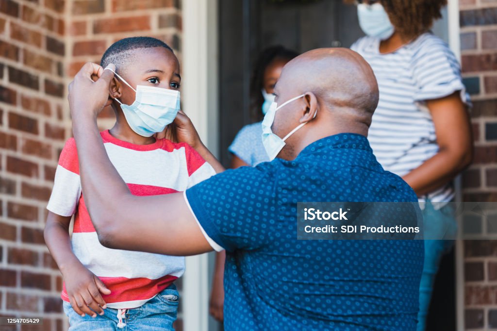 Father helps son put on protective face mask A mid adult father helps his young son put on a protective face mask before leaving their home amid the coronavirus pandemic. Protective Face Mask Stock Photo