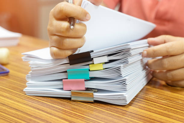 istock Teacher hand holds pen for checking student's homework assignment on table office. Paper documents stacked in archive with paperclip. Report papers stacks. Business and education concept. 1254705483