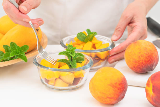 mangues hachées et pêches dans un bol en verre avec des feuilles de menthe de près sur la table de cuisine, les mains des femmes. salade de fruits frais, délicieux dessert, petit déjeuner - fruit vegetable fruit bowl peaches photos et images de collection