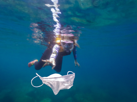 Volunteer girl snorkeling with swimsuit and picking up a mask garbage on the ocean. Cleaner collecting trash on the beach. Tidying up rubbish. Volunteering and recycling coronavirus concept.