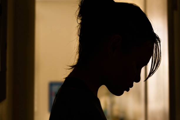 Depressed sad woman at home alone. A woman standing in a shadow hallway of her home looking down in saddness and misery.  Silhoutte. sad girl crouching stock pictures, royalty-free photos & images