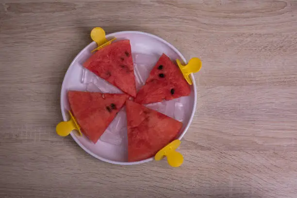 Photo of Watermelon slices in a bowl with ice on an ice cream stick on a wooden background. Creative idea