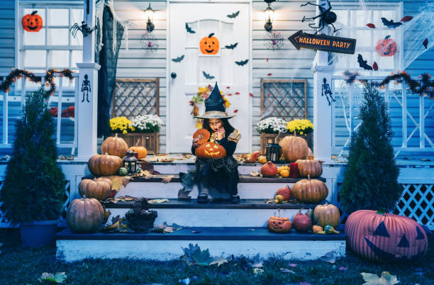魔女の衣装を着た小さな女の子が家の前の階段に座り、ハロウィーンのトリックや御馳走でジャック・オー・ランタン・パンプキンズを抱いている - candy house ストックフォトと画像