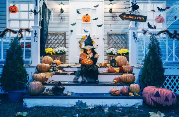 Photo of Little girl in witch costume sitting on the stairs in front of the house and holding Jack-o-Lantern Pumpkins on Halloween trick or treat