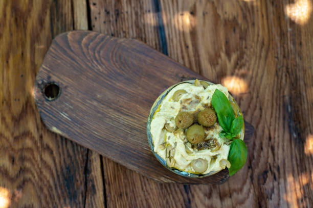 Classic appetizer hummus with olives and a basil branch of chickpea, tahini, olive oil, and oriental spices in a bowl on a wooden table. Traditional Mediterranean vegetarian snack.