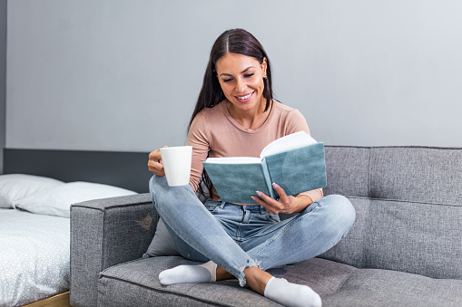 Young woman at home sitting on modern sofa relaxing in her living room reading book and drinking coffee or tea. Cozy bed and a beautiful girl, reading a book, concepts of home and comfort