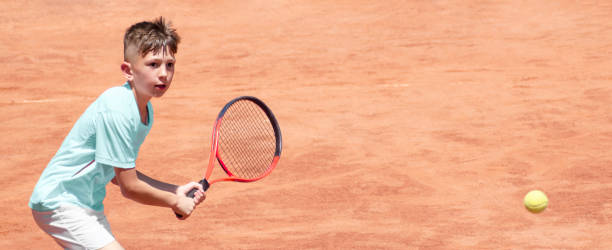 Child on tennis court with a racket in his hands is ready to hit backhand. Boy tennis player in motion and action. Emotions on face of young athlete during sports game. Panoramic. Banner. Copy space niño en la cancha de tenis con una raqueta en sus manos está listo para golpear el revés. jugador de tenis chico en movimiento y acción. emociones en la cara del joven atleta durante el juego deportivo. panorámica. bandera. copiar espacio
