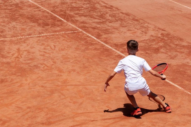 jovem tenista masculino com raquete em ação. o garoto joga tênis em uma quadra de tênis de barro. criança está concentrada e focada no jogo. fundo esportivo de tênis infantil com sombra. movimento. copiar espaço - tennis child childhood sport - fotografias e filmes do acervo