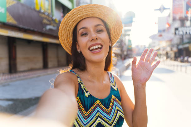 Young brunette woman using a mobile phone takes a self video selfie with attractive smile. Young brunette woman using a mobile phone takes a selfie with attractive smile. Happy Hispanic girl traveler wear summer dress with hat waving to internet audience. Influencer and vlogging concept beautiful mexican girls stock pictures, royalty-free photos & images