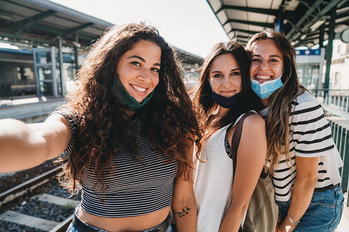 Three friends taking a selfie together - Pov view. They are in a train station, wearing protective facemasks.