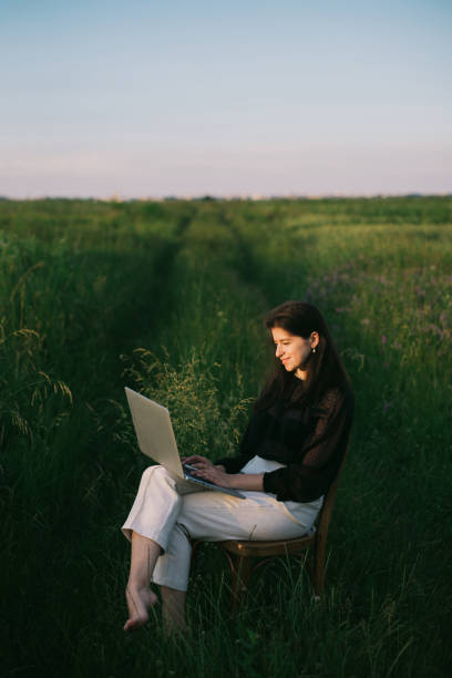 Young business woman working online. Freelance and remote work outdoors. Creative image. Fashionable elegant girl working on laptop and sitting on rustic chair  in summer field at sunset.