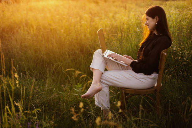 istock Young business woman working online. Freelance and remote work outdoors. Creative image. Fashionable elegant girl with laptop sitting on rustic chair in sunny summer field at sunset. 1254769705