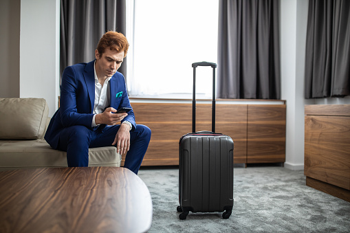 Redhead businessman sitting on sofa and text messaging at hotel suite, suitcase is next to him