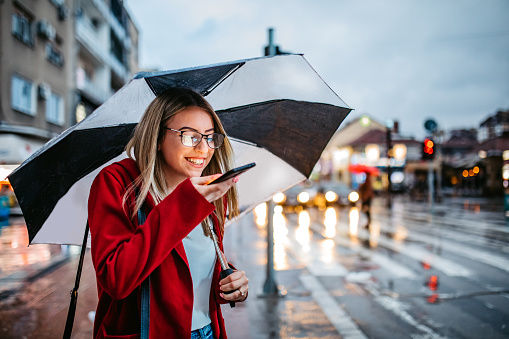 Young Caucasian beautiful smiling woman leaving voice message on smartphone in the rain.
