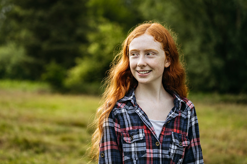 Partial front view of relaxed and happy redhead Caucasian girl wearing casual plaid shirt and looking away from camera in green meadow.