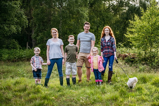 Full length front view of Caucasian parents in mid 30s with children aged 5-15 and small dog standing together in woodland meadow and smiling at camera.