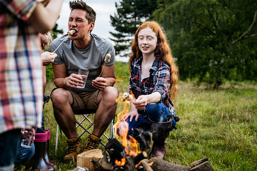 Redhead teenage daughter roasting marshmallows with mid adult father and family on camping trip in late spring.