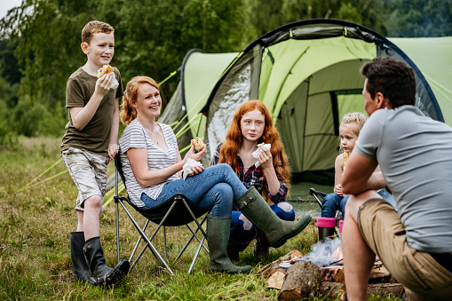 Caucasian family with three children aged 5-15 smiling as they eat meal grilled over open fire while camping in woodland area.
