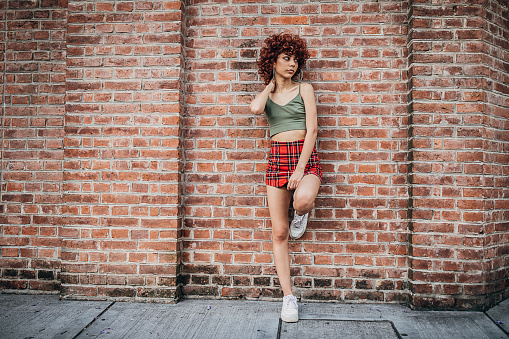 One young woman with red curly hair posing by the brick wall on the street.