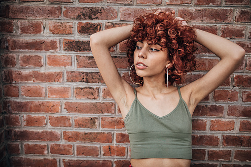 One young woman with red curly hair posing by the brick wall on the street.