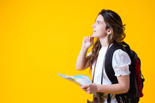 Beautiful young woman tourist with backpack and camera,holding map in hand and looking up copy space for your advertising text.Travel brunette girl thinking about route,isolated on yellow background.