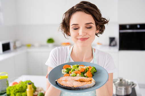 Close up photo of pretty housewife lady chef hold ready grilled, salmon trout fillet steak with garnish cook dinner one person portion eyes closed wear apron t-shirt modern kitchen indoors